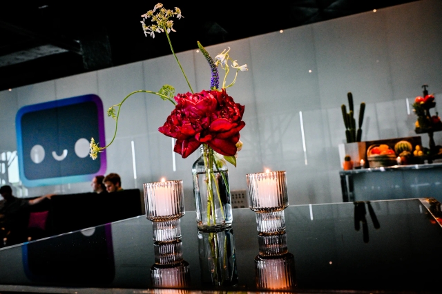 A rose floral in a small vase with water in between two candles on a high black table. In the background is a taco bar to the right and a lounge with a large Houston decal to the left.