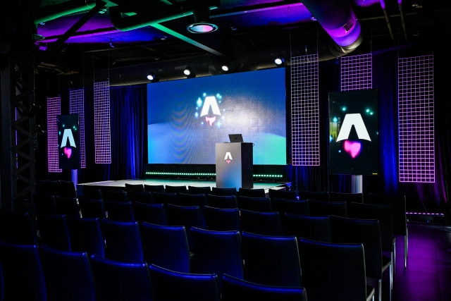 View from the back of the dark empty keynote presentation area. Rows empty black chairs. A vibrant large LED screen with the Astro Together logo on it. Two smaller portrait-oriented screens adorn the sides of the stage, also with the Astro Together logo.