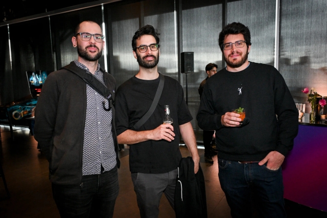 Three men in dark clothing and glasses pose together with drinks.