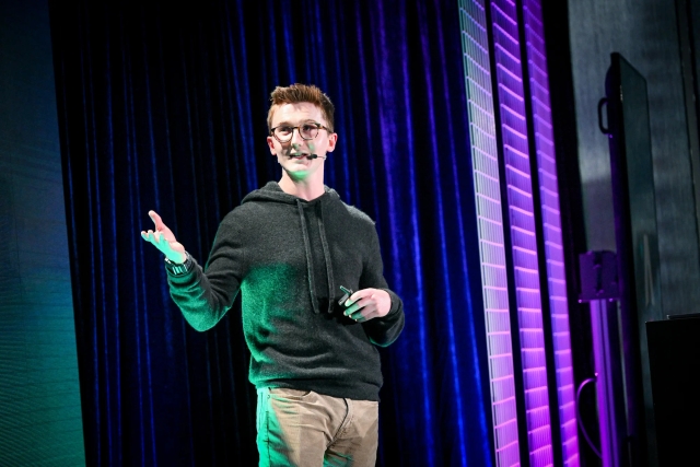 A close up of Ben Holmes on stage gesturing to and looking at the LED screen.