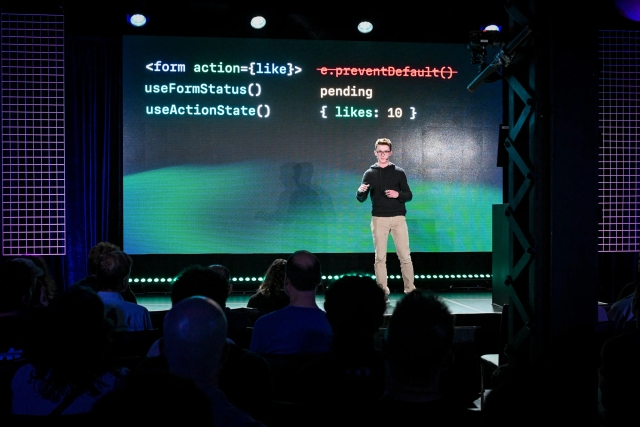 Ben Holmes on stage in front of a slide depicting code for a form with a like button.