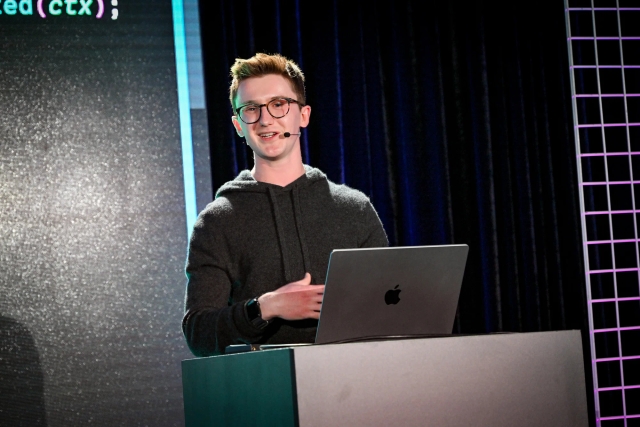 Ben Hlmes on stage at the podium, smiling at the audience.