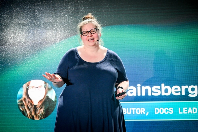 Sarah Rainsberger on stage, introducing herself. Her right hand gestures towards the audience while the left holds a clicker.